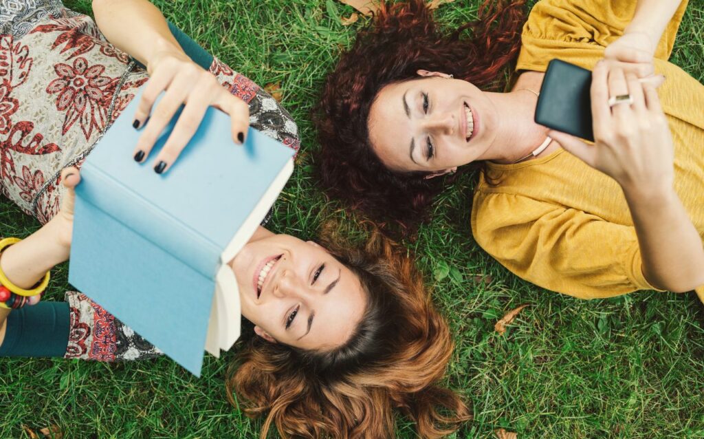 dua orang wanita sedang tidur di atas rumput sambil main hp dan membaca buku tentang ide kerja part time untuk mahasiswa