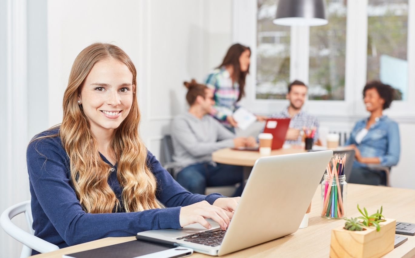 seorang wanita karyawan magang memakai baju kaos biru lengan panjang sedang duduk di depan laptop warna silver dan tersenyum ke arah kamera