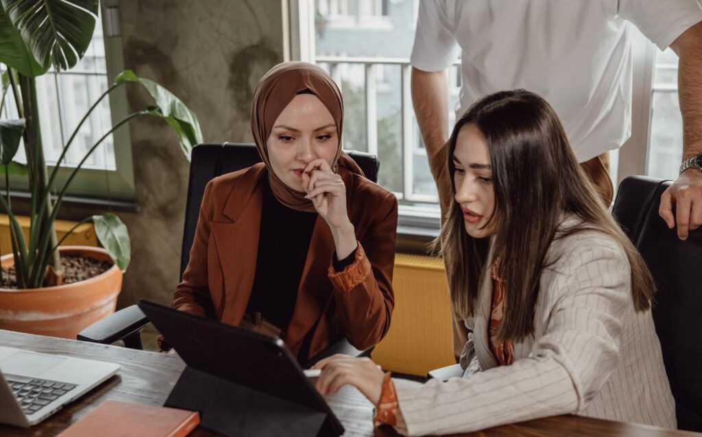 seorang wanita menggunakan hijab berwarna bata dan seorang wanita dengan rambut coklat berkilau sedang bersama-sama melihat ke arah ipad hitam dengan muka bingung karena ingin tau cara menghindari lowongan kerja yang palsu
